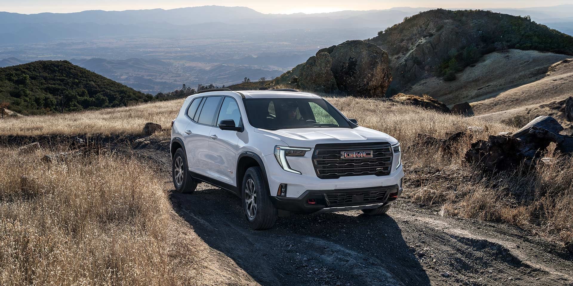 White GMC Acadia SUV parked on a rugged mountain trail with a scenic landscape backdrop.